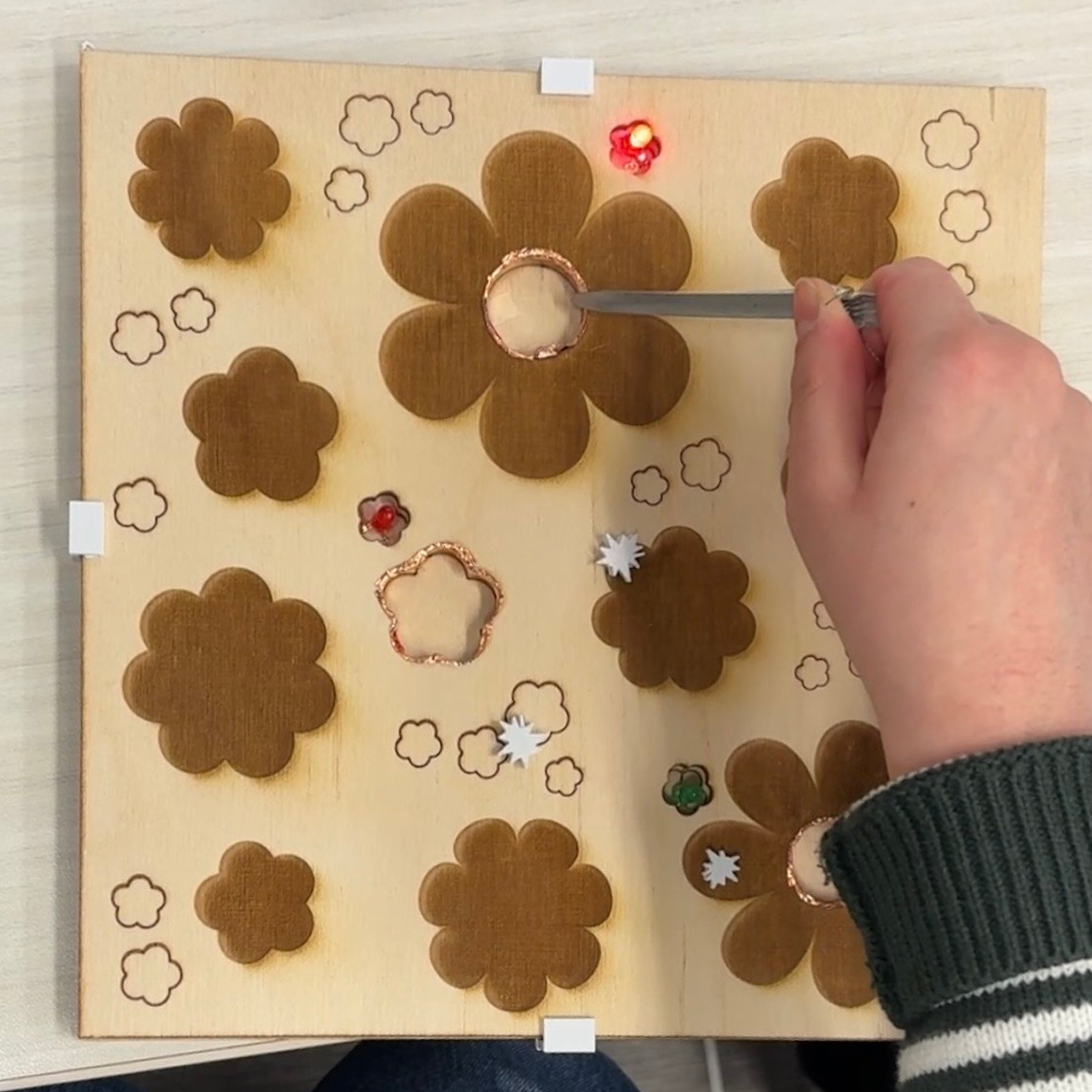 This non-wearable uses multiple simple circuits (the three holes) that are all connected to one switch (the metal tweezers) which can be used to activate any of them upon touching the copper tape lined on the edges of each hole. The project itself was designed by me then laser cut to fit the overall aesthetic of the game.